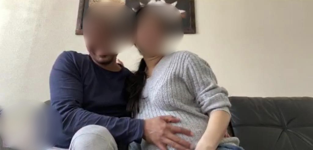 A couple with their faces blurred look at the camera. The man rests his hand sweetly on the woman's heavily pregnant stomach
